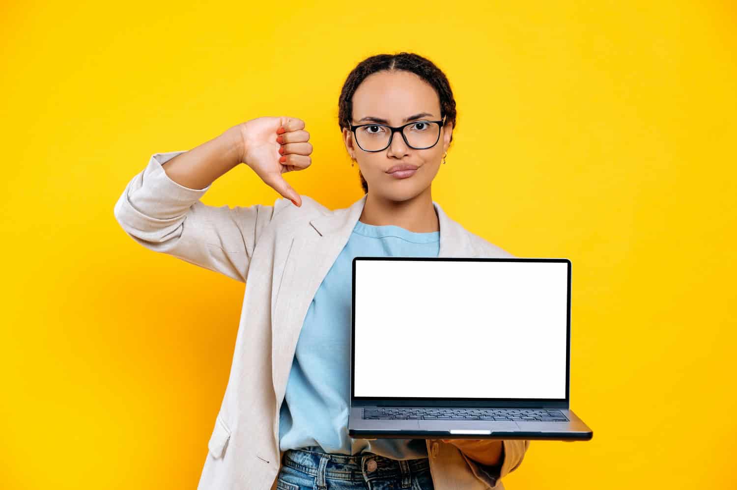Confused young brunette mixed race woman, hold laptop computer with blank mock-up copy space screen, stand on isolated orange background, looks at camera, shows thumb down, dislike gesture